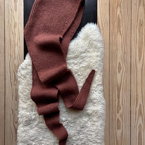 K&ouml;nnte beinhalten: Ein brauner gestrickter Kapuzen-Schal mit einem langen Schwanz. Der Schal liegt auf einer wei&szlig;en, flauschigen Oberfl&auml;che.