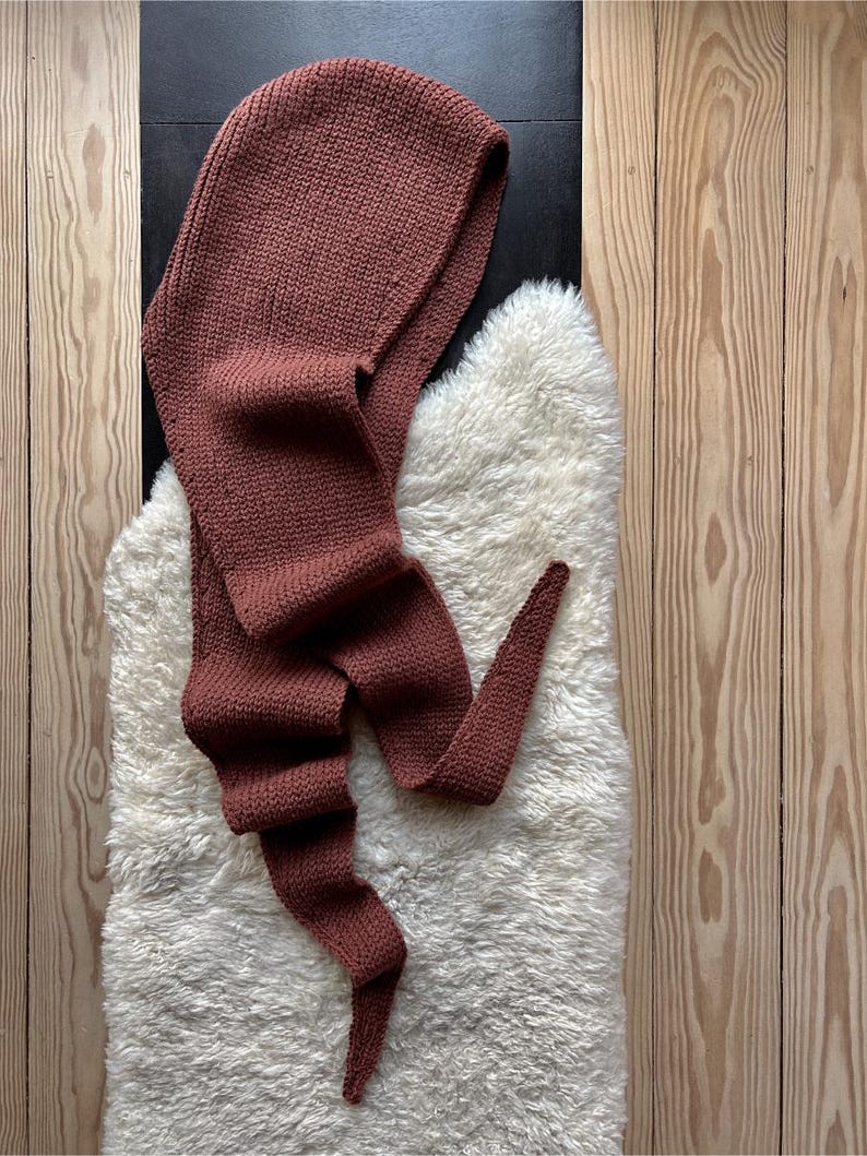 K&ouml;nnte beinhalten: Ein brauner gestrickter Kapuzen-Schal mit einem langen Schwanz. Der Schal liegt auf einer wei&szlig;en, flauschigen Oberfl&auml;che.