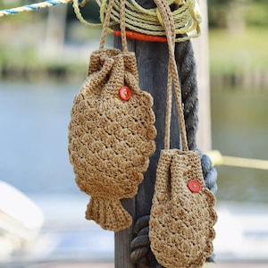 May include: Two handmade, fish-shaped crochet bags in a natural tan color, each with a red button closure. The bags hang from a wooden post with rope and nautical elements in the background. The bags have a textured, scale-like pattern.