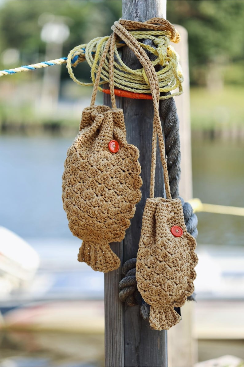 May include: Two handmade, fish-shaped crochet bags in a natural tan color, each with a red button closure. The bags hang from a wooden post with rope and nautical elements in the background. The bags have a textured, scale-like pattern.