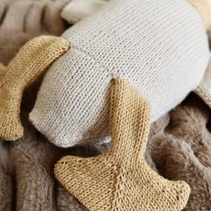 May include: A knitted plush toy in the shape of a goose, with a cream-colored body and tan-colored feet and beak. The toy is lying on a brown, textured surface.