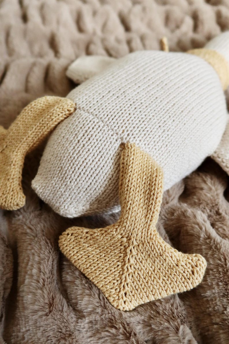 May include: A knitted plush toy in the shape of a goose, with a cream-colored body and tan-colored feet and beak. The toy is lying on a brown, textured surface.