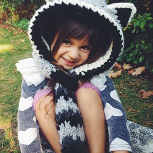 May include: A young girl wearing a gray and black crocheted raccoon hat with a matching scarf. The hat has large ears and a white crocheted trim. The girl is wearing a gray sweater with white hearts and a black and white striped scarf.