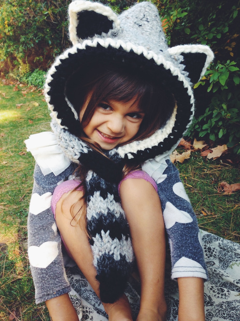 May include: A young girl wearing a gray and black crocheted raccoon hat with a matching scarf. The hat has large ears and a white crocheted trim. The girl is wearing a gray sweater with white hearts and a black and white striped scarf.