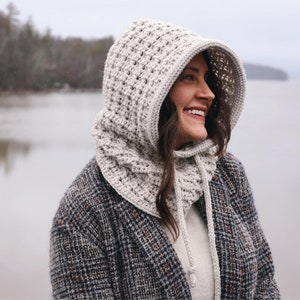 May include: A white crocheted hooded cowl with a long, flowing design. The cowl is worn by a person standing in front of a lake.
