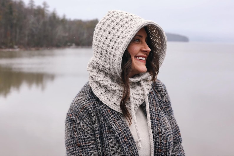 May include: A white crocheted hooded cowl with a long, flowing design. The cowl is worn by a person standing in front of a lake.