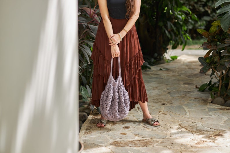 May include: A woman wearing a brown maxi skirt with ruffles and a crocheted lavender tote bag.