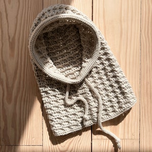 May include: A beige crocheted hooded scarf with a long tie. The scarf is lying on a wooden surface.