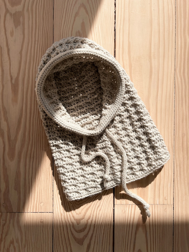 May include: A beige crocheted hooded scarf with a long tie. The scarf is lying on a wooden surface.