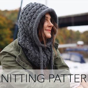 May include: A gray knitted hooded cowl with a cable stitch pattern. The cowl is worn by a person in a green jacket, standing outdoors.