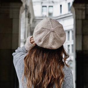 Puede incluir: Una mujer con un gorro marr&oacute;n claro y un su&eacute;ter gris. Est&aacute; de pie en una puerta con un edificio en el fondo.