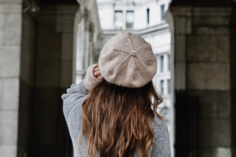 Puede incluir: Una mujer con un gorro marr&oacute;n claro y un su&eacute;ter gris. Est&aacute; de pie en una puerta con un edificio en el fondo.