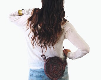 Può includere: Una borsa rotonda di uncinetto marrone e viola con una lunga tracolla indossata sulla spalla. La borsa è indossata da una persona che indossa una camicia bianca a maniche lunghe e pantaloncini di jeans. CROCHET PATTERN è scritto in nero su sfondo bianco.