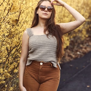 Peut inclure: Une femme portant un d&eacute;bardeur en crochet marron clair avec un d&eacute;collet&eacute; carr&eacute; et un pantalon marron. Elle se tient devant un buisson avec des fleurs jaunes.