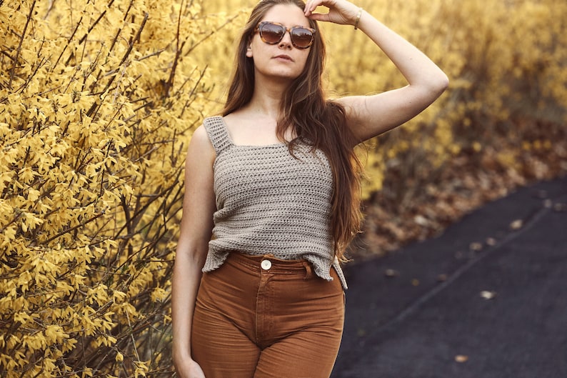 Peut inclure: Une femme portant un d&eacute;bardeur en crochet marron clair avec un d&eacute;collet&eacute; carr&eacute; et un pantalon marron. Elle se tient devant un buisson avec des fleurs jaunes.