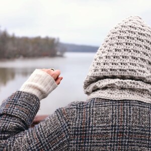 May include: A person wearing a gray and white hooded sweater with a plaid jacket, standing by a lake with trees in the background.