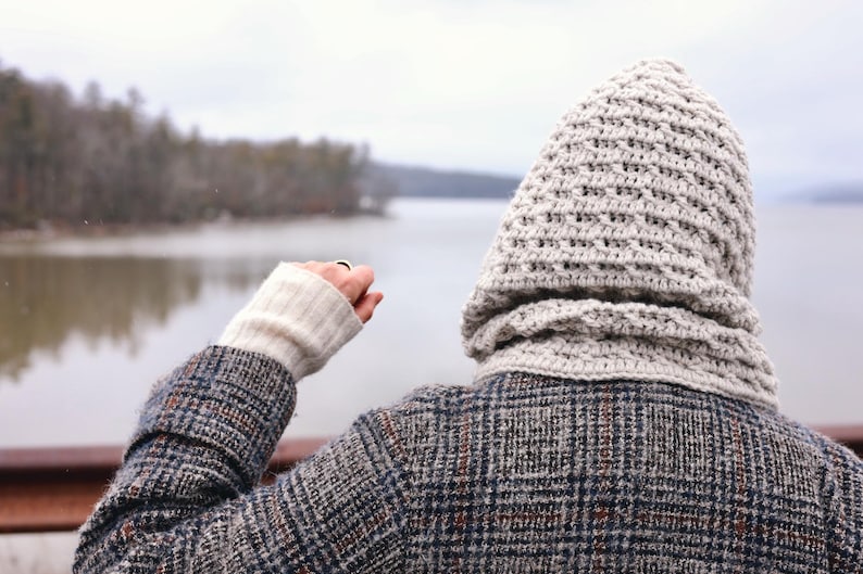 May include: A person wearing a gray and white hooded sweater with a plaid jacket, standing by a lake with trees in the background.