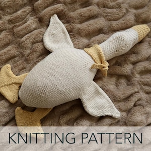 以下が含まれることがあります： クリーム色と淡い黄色で編まれたガチョウ型の枕。ガチョウには翼、足、ネクタイが付いています。画像の下部には「KNITTING PATTERN」という文字があります。