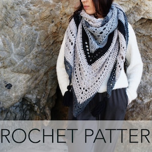 May include: A crochet shawl in black, grey and white, with a triangular shape and fringe border. The shawl is being worn by a person standing in front of a rocky background.