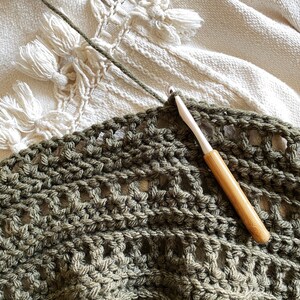 May include: A close-up of a crocheted blanket in a dark green color. The blanket is being worked on with a wooden crochet hook.