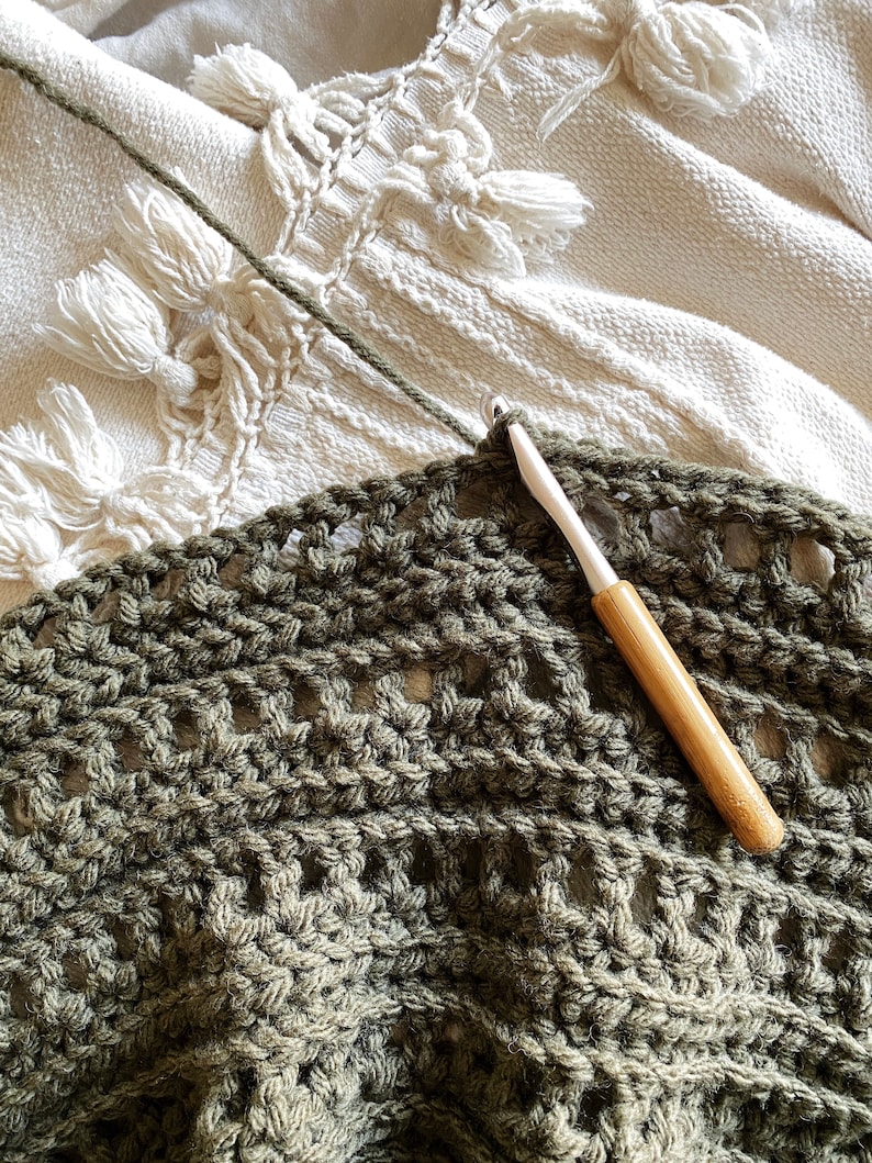 May include: A close-up of a crocheted blanket in a dark green color. The blanket is being worked on with a wooden crochet hook.