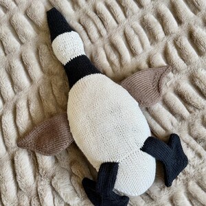 May include: A knitted goose plush toy in cream, black, and brown. The goose has a black head and feet, cream-colored body, and brown wings. The toy is lying on a textured, light brown surface.