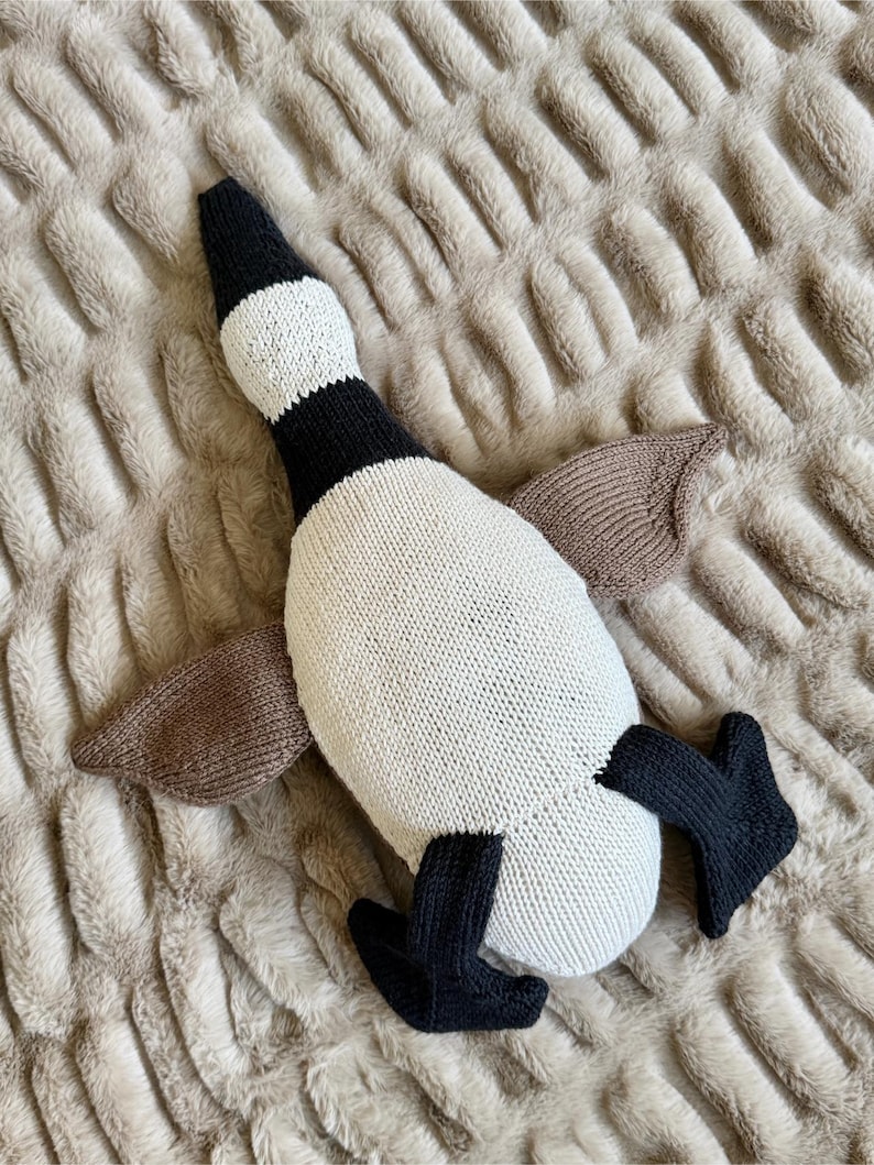 May include: A knitted goose plush toy in cream, black, and brown. The goose has a black head and feet, cream-colored body, and brown wings. The toy is lying on a textured, light brown surface.