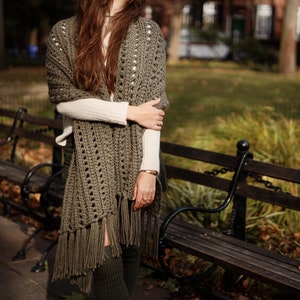 May include: A woman wearing a green crochet shawl with fringe. The shawl is draped over her shoulders and tied at her neck. She is standing in front of a wooden bench.