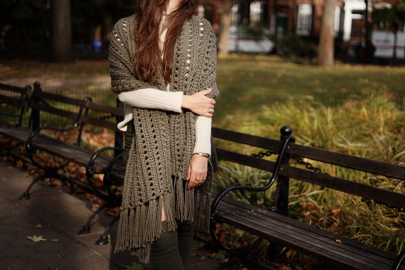 May include: A woman wearing a green crochet shawl with fringe. The shawl is draped over her shoulders and tied at her neck. She is standing in front of a wooden bench.