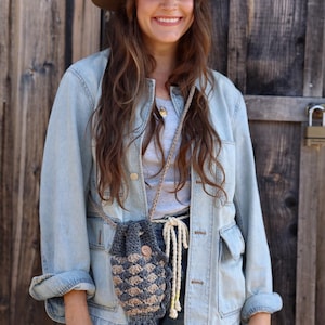 May include: A crocheted crossbody bag in shades of gray and beige, featuring a textured pattern and a wooden button closure. The bag is suspended by a beige rope strap. The person is wearing a denim jacket and a brown hat.