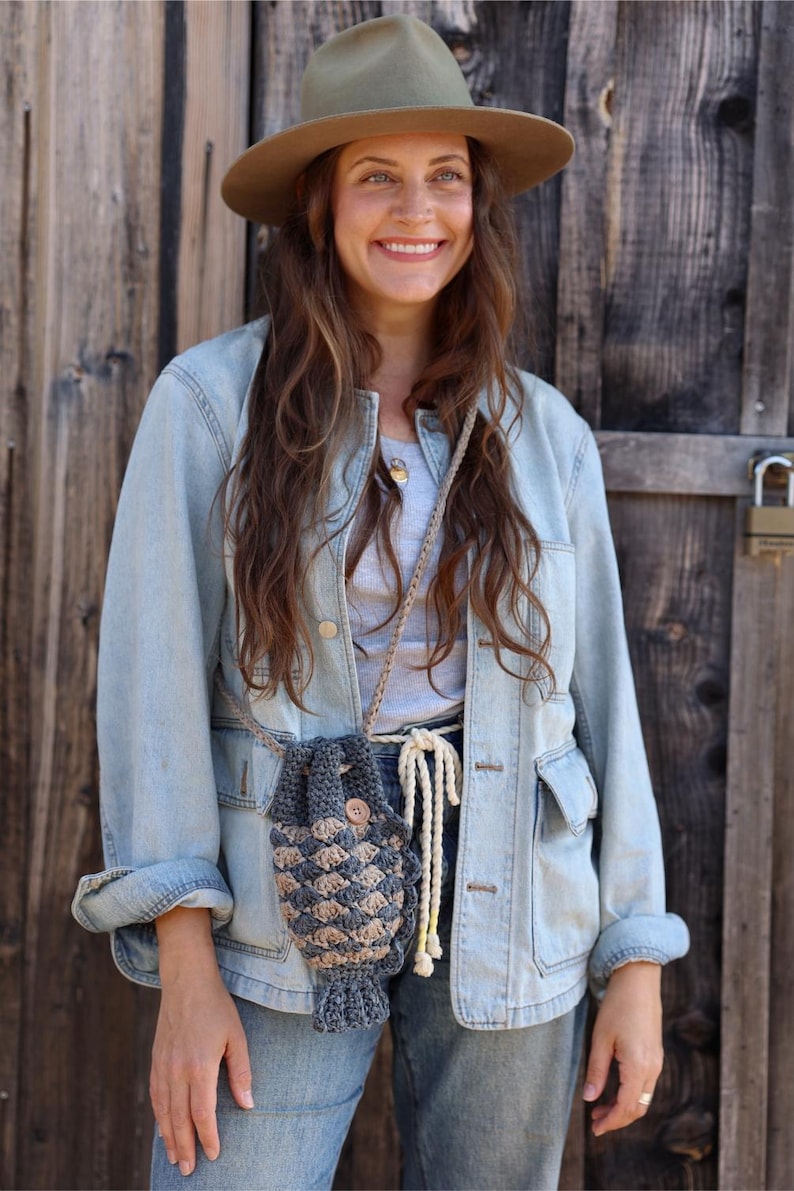 May include: A crocheted crossbody bag in shades of gray and beige, featuring a textured pattern and a wooden button closure. The bag is suspended by a beige rope strap. The person is wearing a denim jacket and a brown hat.