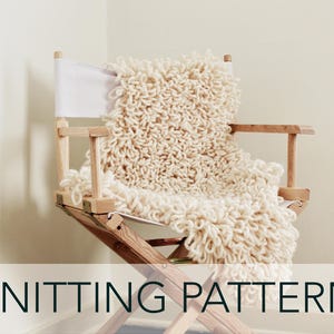 Puede incluir: Una manta de punto blanca drapeada sobre una silla de director de madera. La manta tiene un aspecto esponjoso y texturizado.  KNITTING PATTERN está escrito en negro en la parte inferior de la imagen.
