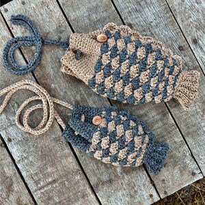 May include: Two handmade fish-shaped bags with long straps. The bags are crocheted with a tan and blue diamond pattern, featuring wooden button closures. The bags are lying on a weathered wooden surface.