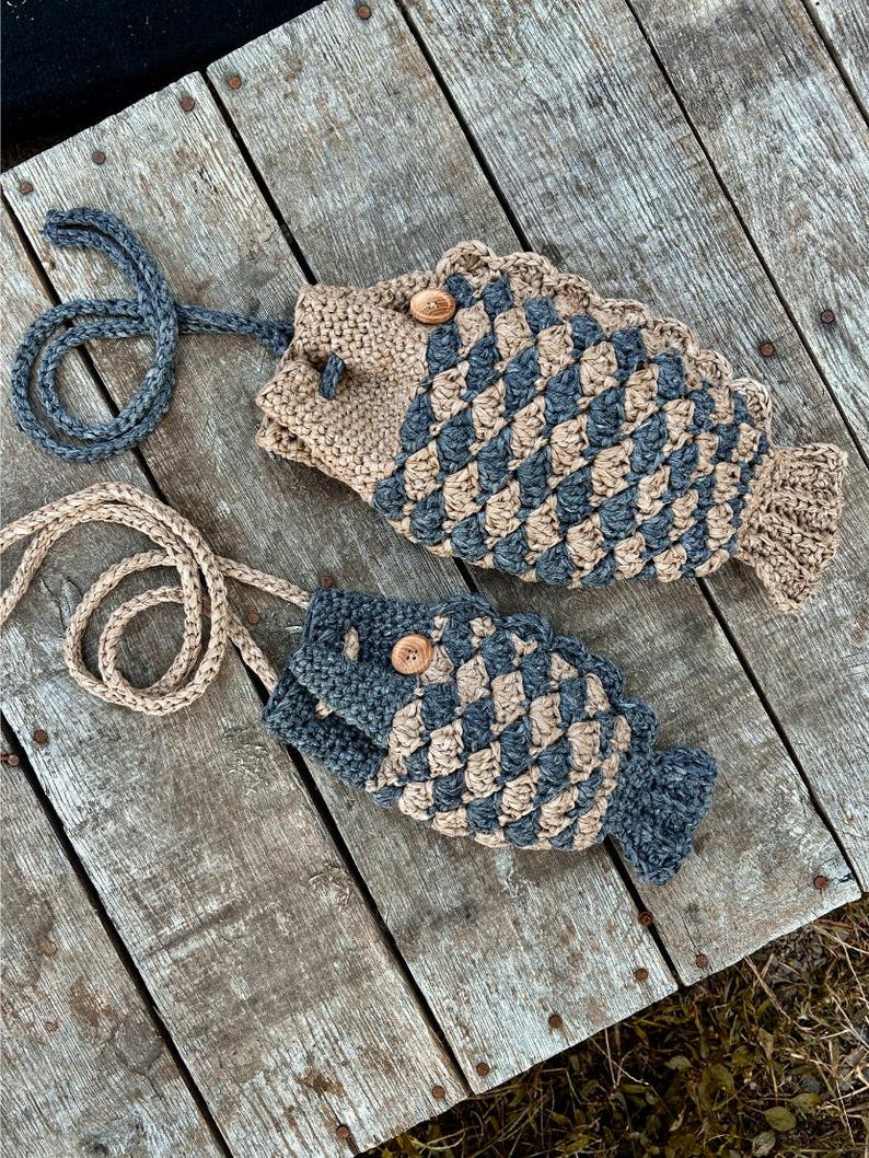 May include: Two handmade fish-shaped bags with long straps. The bags are crocheted with a tan and blue diamond pattern, featuring wooden button closures. The bags are lying on a weathered wooden surface.