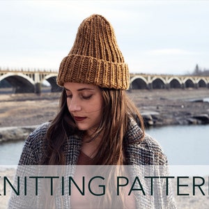 May include: A brown knitted beanie hat with a ribbed texture. The hat is worn by a person standing in front of a bridge and a river.