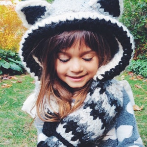 May include: A young person wearing a gray and black crocheted raccoon hat with a matching scarf. The hat has large ears and a pointed hood.