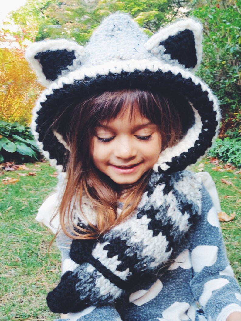May include: A young person wearing a gray and black crocheted raccoon hat with a matching scarf. The hat has large ears and a pointed hood.