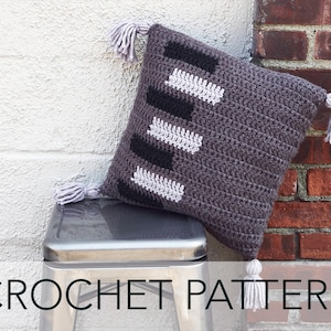 May include: A gray crochet pillow with black and white geometric patterns and tassels.  "CROCHET PATTERN" is written in black text at the bottom of the image.