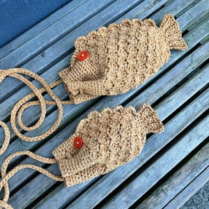 May include: Two handmade, fish-shaped crochet bags in a light brown color, each featuring a red button eye and a rope strap. The bags are lying on a weathered wooden bench, showcasing their unique design and texture.