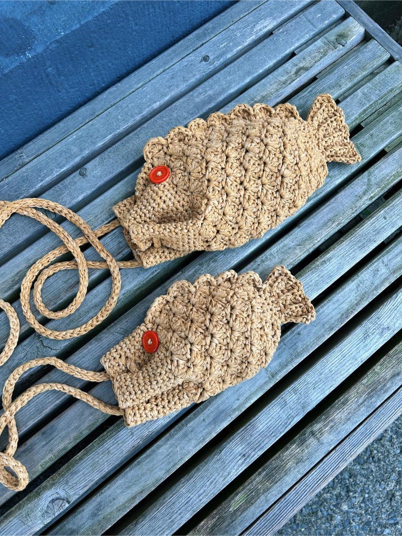 May include: Two handmade, fish-shaped crochet bags in a light brown color, each featuring a red button eye and a rope strap. The bags are lying on a weathered wooden bench, showcasing their unique design and texture.