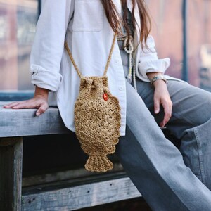 May include: A tan crochet fish-shaped crossbody bag with a drawstring closure and a small red button. The bag has a textured pattern and a long, thin strap. The bag is approximately 10 inches long.