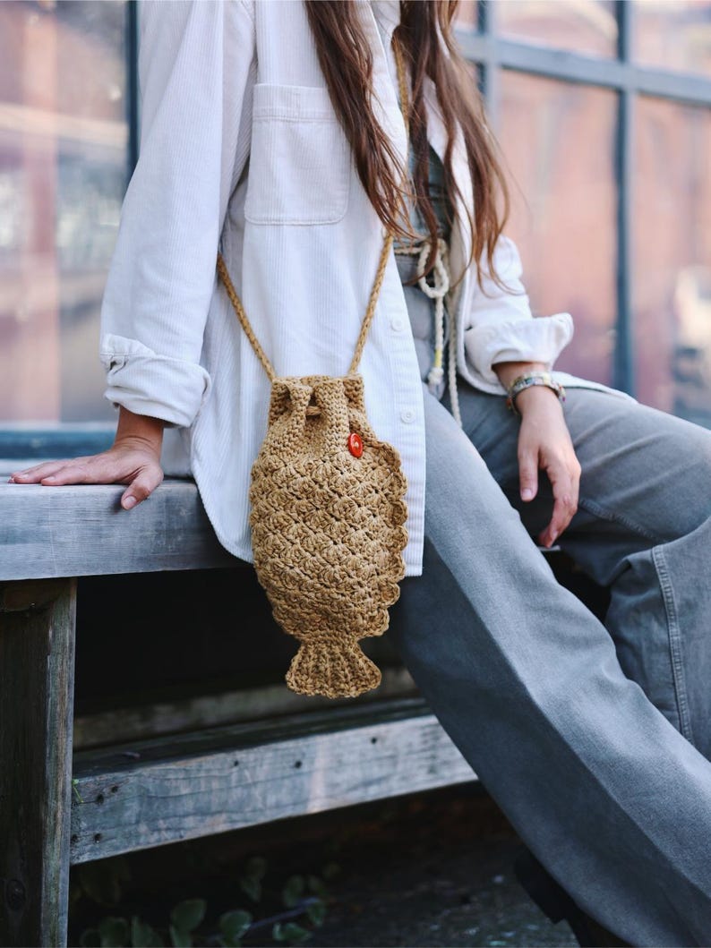 May include: A tan crochet fish-shaped crossbody bag with a drawstring closure and a small red button. The bag has a textured pattern and a long, thin strap. The bag is approximately 10 inches long.