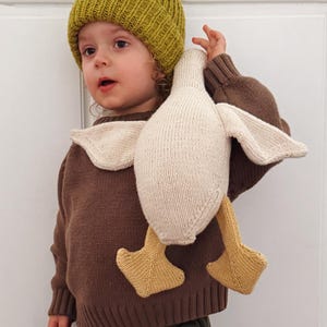 May include: A child wearing a green knit beanie and a brown sweater holds a knitted duck toy. The duck has cream-colored wings and a yellow beak and feet. The child is also wearing olive green pants.