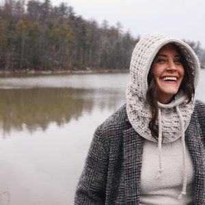 May include: A woman wearing a white crocheted hooded cowl with a gray and black plaid jacket. She is standing in front of a lake with trees in the background.