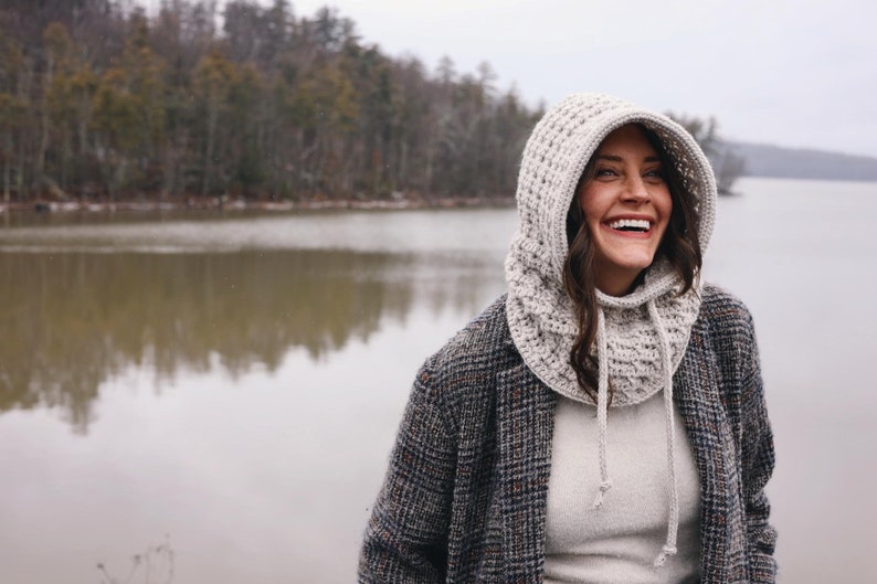 May include: A woman wearing a white crocheted hooded cowl with a gray and black plaid jacket. She is standing in front of a lake with trees in the background.