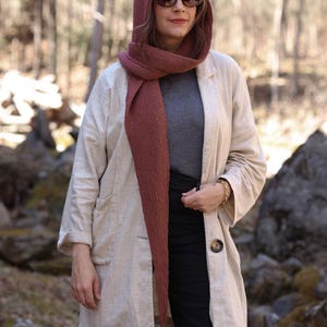 K&ouml;nnte beinhalten: Eine Frau tr&auml;gt eine lange, beige Leinenjacke mit einem gro&szlig;en Knopfverschluss. Sie tr&auml;gt au&szlig;erdem einen langen, burgunderfarbenen Strick Schal und einen passenden Schal mit Kapuze. Sie tr&auml;gt eine Sonnenbrille und hat lange braune Haare.