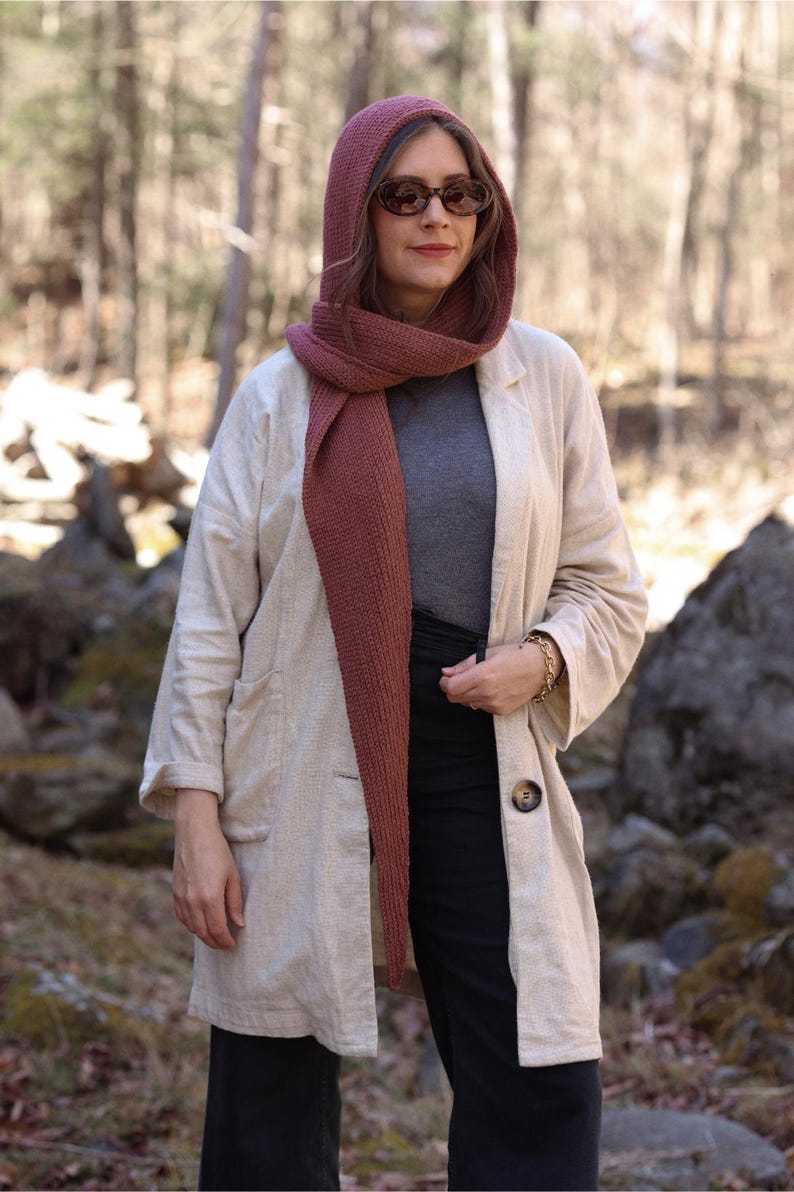 K&ouml;nnte beinhalten: Eine Frau tr&auml;gt eine lange, beige Leinenjacke mit einem gro&szlig;en Knopfverschluss. Sie tr&auml;gt au&szlig;erdem einen langen, burgunderfarbenen Strick Schal und einen passenden Schal mit Kapuze. Sie tr&auml;gt eine Sonnenbrille und hat lange braune Haare.