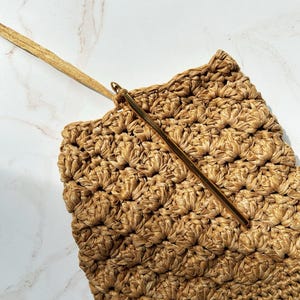 May include: Close-up of a crochet project in progress. The project is a textured, woven bag in shades of tan and gold. A gold crochet hook is inserted into the fabric, with a long, thin gold strap extending from the bag.