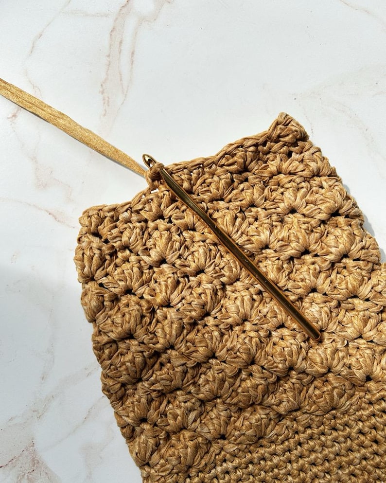 May include: Close-up of a crochet project in progress. The project is a textured, woven bag in shades of tan and gold. A gold crochet hook is inserted into the fabric, with a long, thin gold strap extending from the bag.