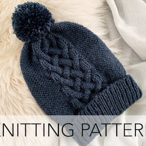 Peut inclure: Un bonnet en tricot bleu foncé avec un gros pompon sur le dessus. Le bonnet est en tricot torsadé et est photographié sur une surface blanche et moelleuse.  KNITTING PATTERN est écrit en blanc en bas de l'image.